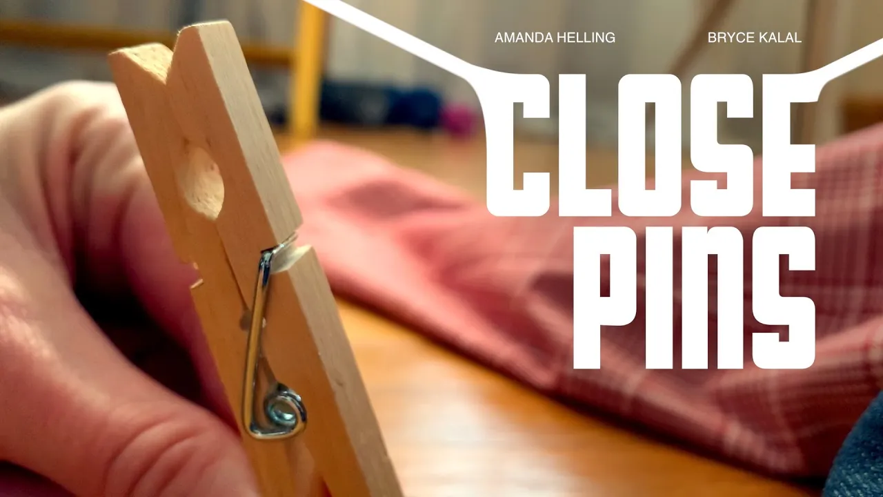 A close-up of a clothespin being held by someone's hand. White text reads "Close Pins. Amanda Helling, Bryce Kalal."