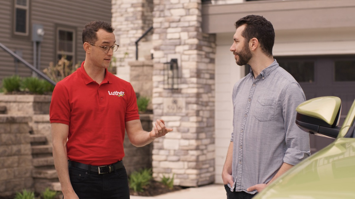 Two men talking on a driveway next to a green car. One is listening with a smile and has his hands in his pocket. The other is wearing a red Luther Auto polo and gesturing toward the car.